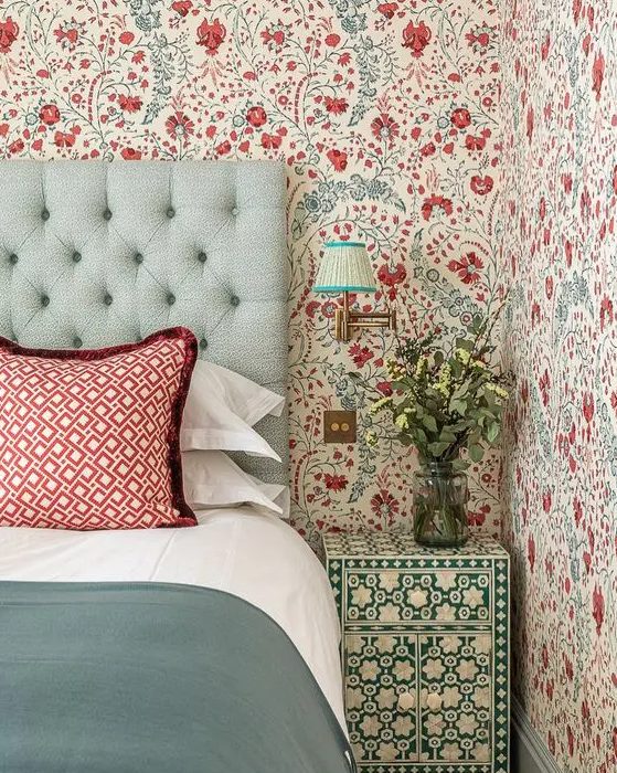 An eye catchy bedroom with bold blue and red floral wallpaper, a mint upholstered bed with red printed and green bedding, a green and white floral nightstand