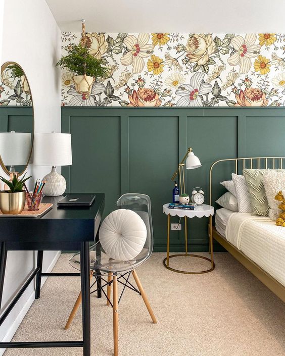 An eye catchy bedroom with bold floral wallpaper, green paneling, a metal bed with neutral bedding, a nightstand, a black vanity and a clear chair