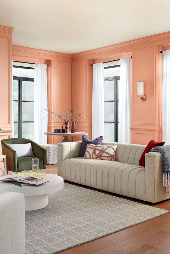 An eye catchy peach pink living room with paneled walls, a grey sofa, a green chair, a coffee table and neutral curtains
