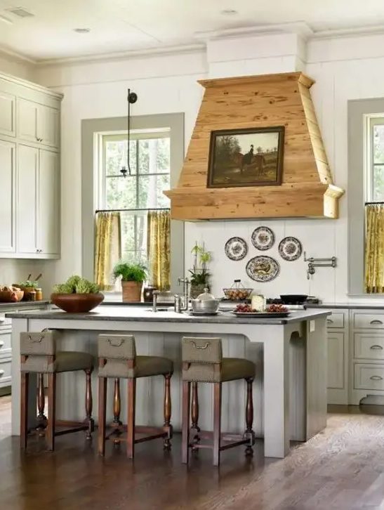 An off white vintage kitchen with shaker cabinets, a stained wood hood with an artwork and yellow curtains