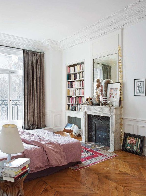 A French chic bedroom with paneling, an antique non working fireplace, a bed with pink bedding, built in shelves and a mirror