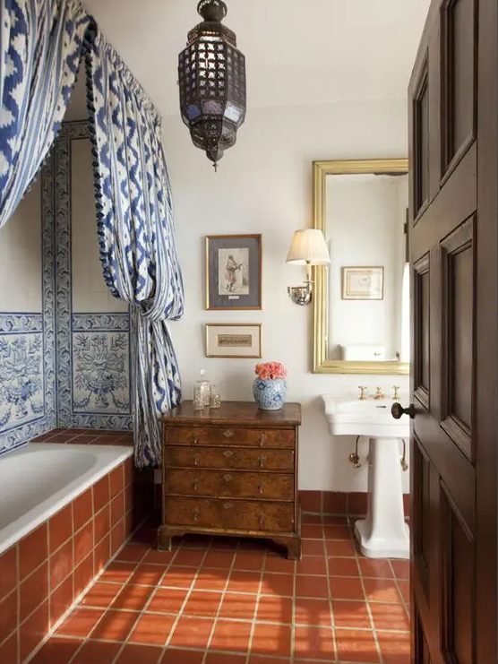 A Moroccan bathroom clad with terracotta tiles, with blue ones and blue curtains, a Moroccan pendant lamp and a dark stained dresser