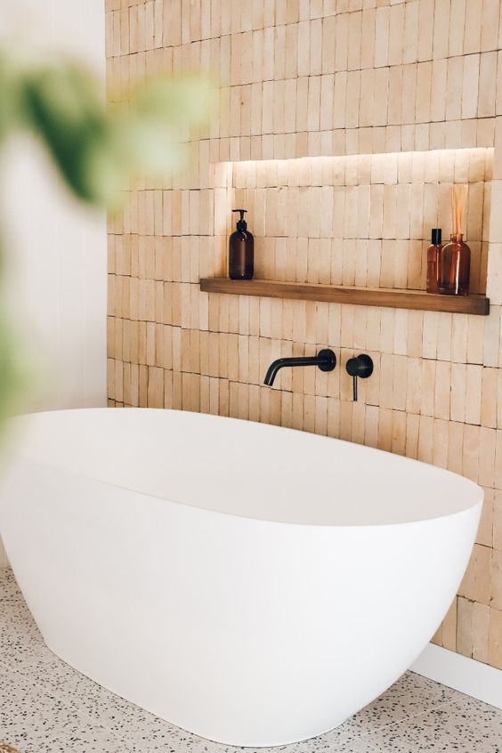 a bathing space clad with terracotta tiles, with a niche and a shelf plus lights, an oval tub and black fixtures