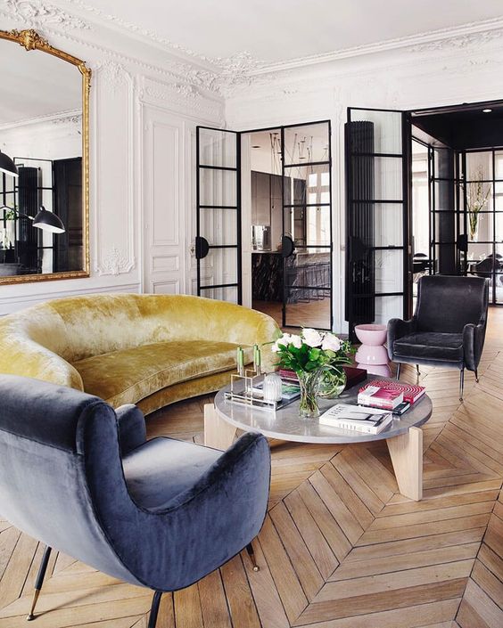 a beautiful Parisian livng room with molding and chevron floors, a mustard sofa, a blue chair, a statement mirror in an ornated frame
