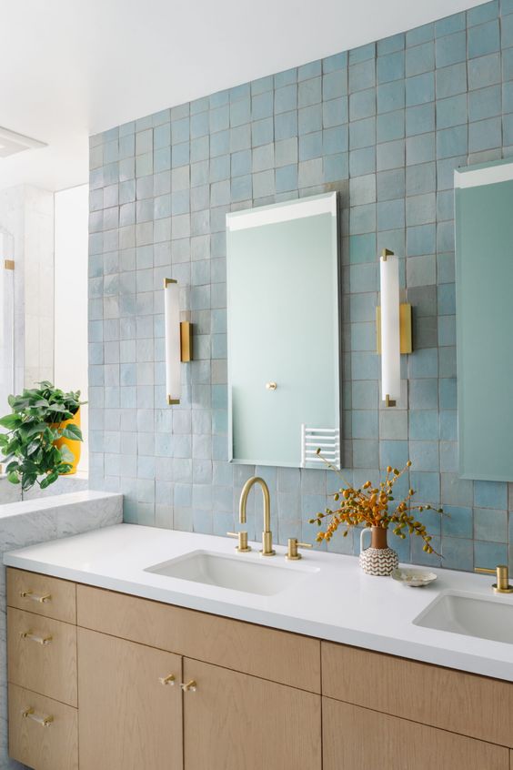 a beautiful bathroom clad with blue Zellige tiles, a light-stained vanity, mirrors and wall sconces plus gold fixtures