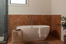 a beautiful bathroom done with terracotta herringbone tiles and blue ones, a tub, a woven side table and rugs