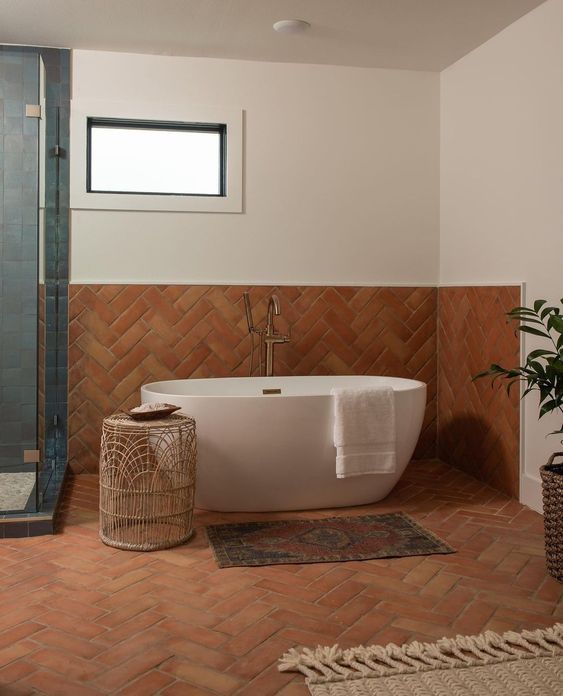 a beautiful bathroom done with terracotta herringbone tiles and blue ones, a tub, a woven side table and rugs