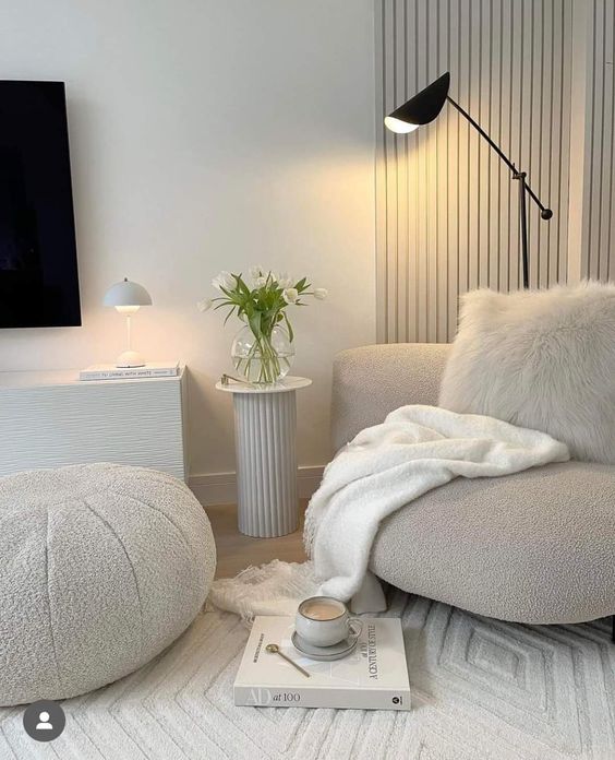 a beautiful neutral nook with a grey boucle chair with a pillow and a blanket, a white boucle pouf, a black lamp, a side table and a geo rug