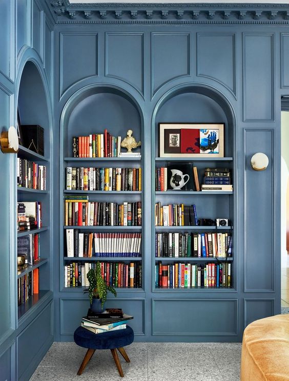 a beautiful space with blue arched bookcases and molding, a navy stool and a yellow seat for reading here