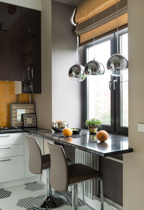 a black and white kitchen with a yellow tile backsplash and a windowsill table plus taupe chair and pendant lamps