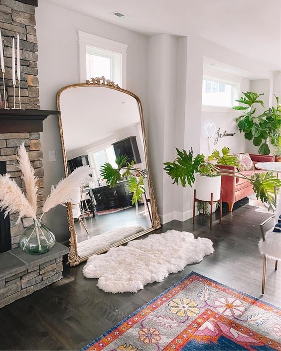 a boho living room with a fireplace clad with stone, an oversized floor mirror, some cool rugs and potted greenery