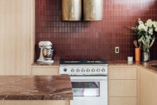 a bold kitchen look with light-stained cabinets, brown stone countertops, a burgundy tile backsplash and gold hood