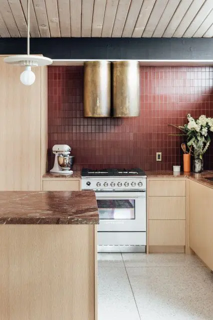 a bold kitchen look with light-stained cabinets, brown stone countertops, a burgundy tile backsplash and gold hood