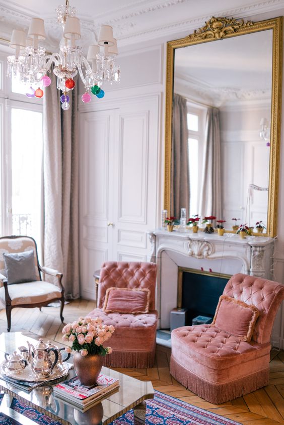 a bright and vivacious space with peachy pink chairs, a fireplace with a large mirror over it, neutral furniture and a chandelier