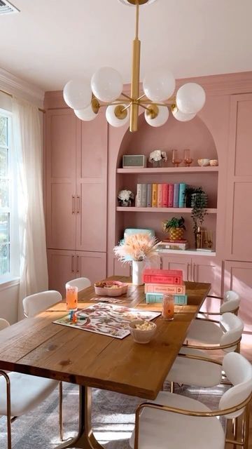 a bright dinign and working space with pink built-in cabinets and an arched bookcase, a stained table and creamy chairs, a chandelier