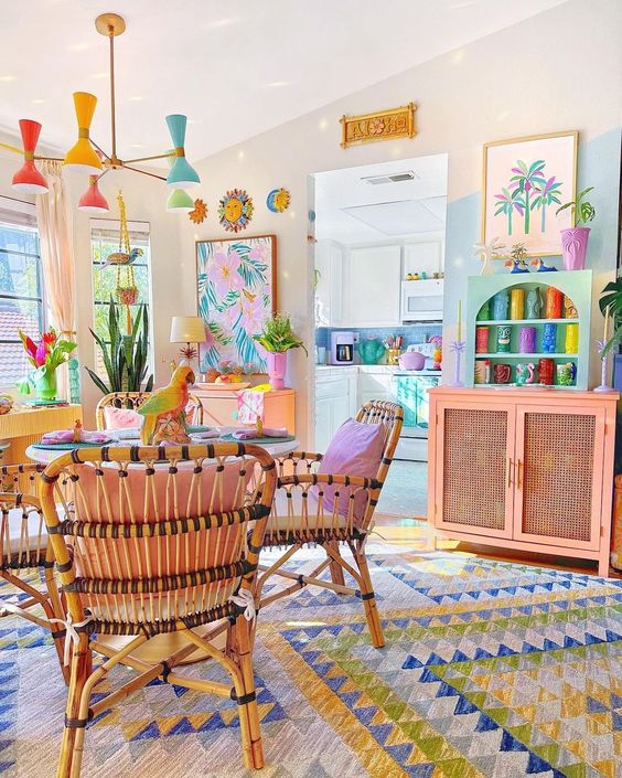 a bright dopamine decor living room with a table and rattan chairs, a bold rug, a pink cabinet and turquoise shelves, a pastel chandelier