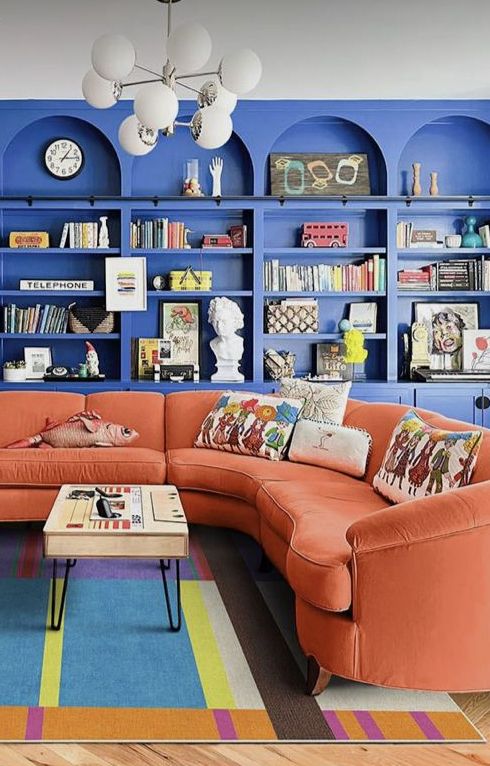 a bright maximalist space with blue arched bookcases, an orange curved sofa, a bright rug, lovely decor and books