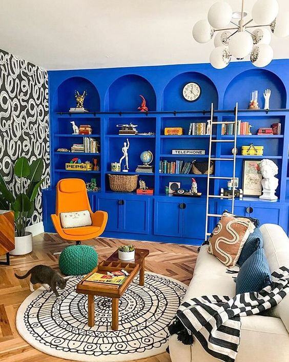 a bright maximalist space with bold blue arched bookcases, a white sofa and pillows, a printed rug, an orange chair and some decor