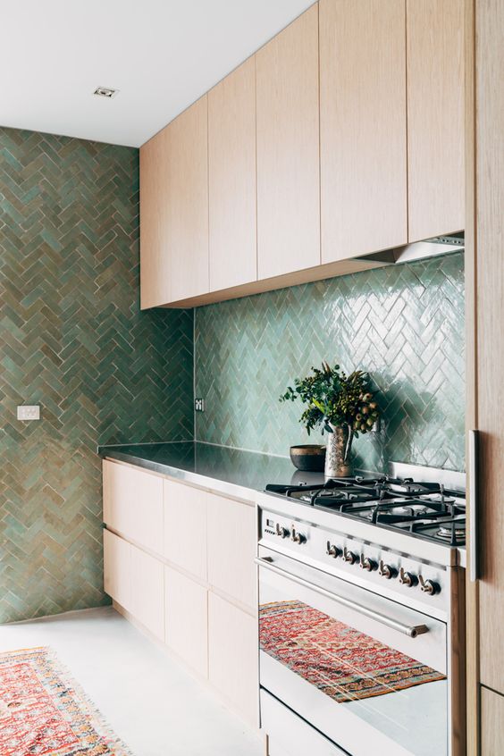 A chic light stained kitchen with sleek cabinets, a green herringbone tile backsplash and a wall, shiny metal countertops and a cooker