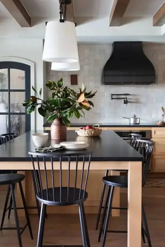 A chic vintage inspired kitchen with stained shaker cabinets and a large table kitchen island, black statement countertops, a black hood and black chairs