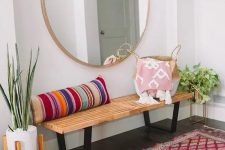 a colorful boho entryway with a wooden bench, a striped pillow, a round mirror, a bright rug and potted plants
