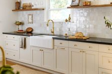 a cozy farmhouse kitchen with tan cabinets, black countertops, a pearly Zellige tile backsplash, stained shelves and gold fixturesHome