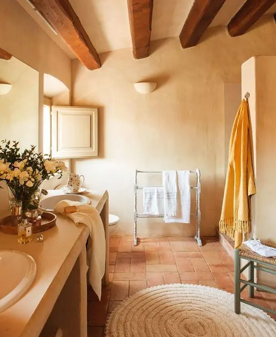 a cozy rustic bathroom with plaster walls and terracotta tiles, a large double vanity, a woven stool and wooden beams on the ceiling