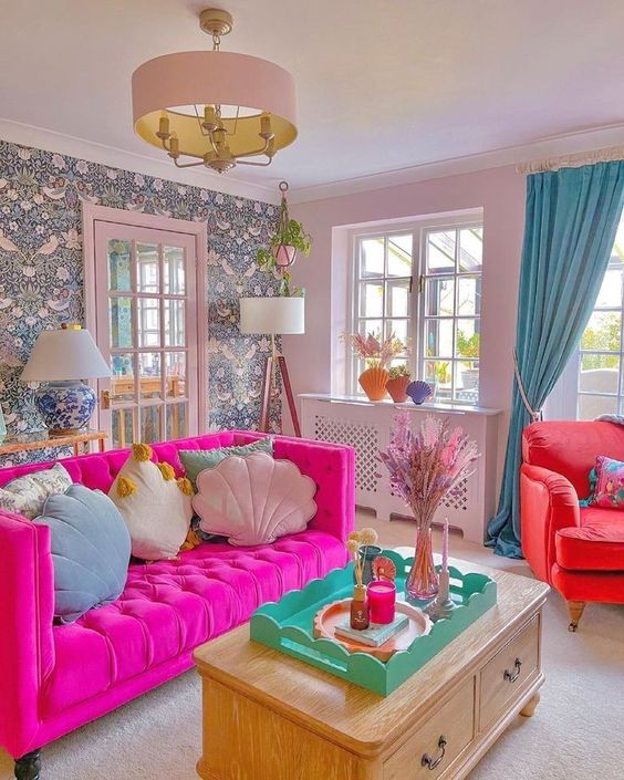a dopamine decor living room with a wallpaper wall, a hot pink sofa with pastel pillows, a coral chair and blue curtains