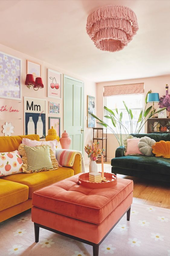 a dopamine decor living room with blush walls, a marigold sofa, a coral ottoman, a dark green chair, a gallery wall and a pink chandelier