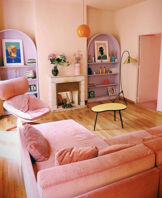 a dopamine decor living room with blush walls and a ceiling, a pink sofa and chair, lilac shelves and a pendant lamp