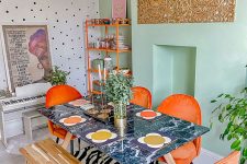 a dopamine dining space with a mint and spotted accent wall, a marble table, orange chairs and a shelving unit, greenery