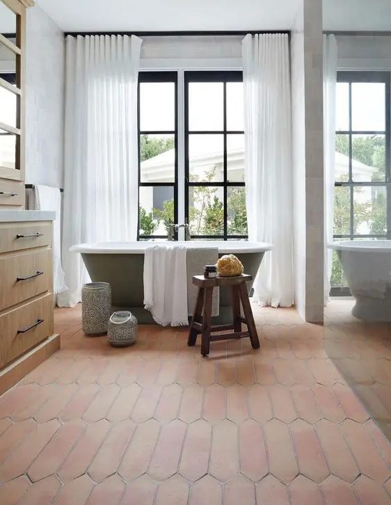 A farmhouse bathroom with a glazed wall, a green bathtub, a stained vanity, a terracotta tile floor and a glass enclosed shower