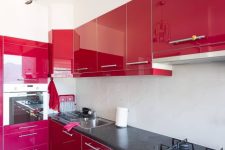 a glossy cherry red kitchen with grey countertops, a sleek white backsplash, a creative chandelier and neutral fixtures