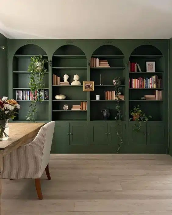 A home office with dark green built in arched bookcases, greenery, a desk and a white chair is a chic space