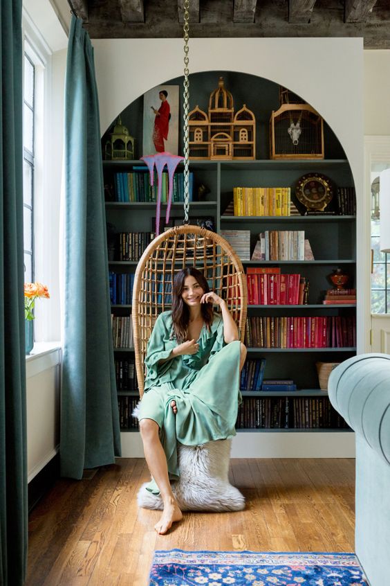 a large arched bookcase with dark green backing and shelves, colorful books and decor, a pendant chair to form a reading nook
