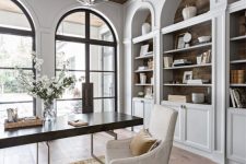 a large home office with arched windows and arched bookcases that echo with them, a desk, a white chair and a sphere lamp