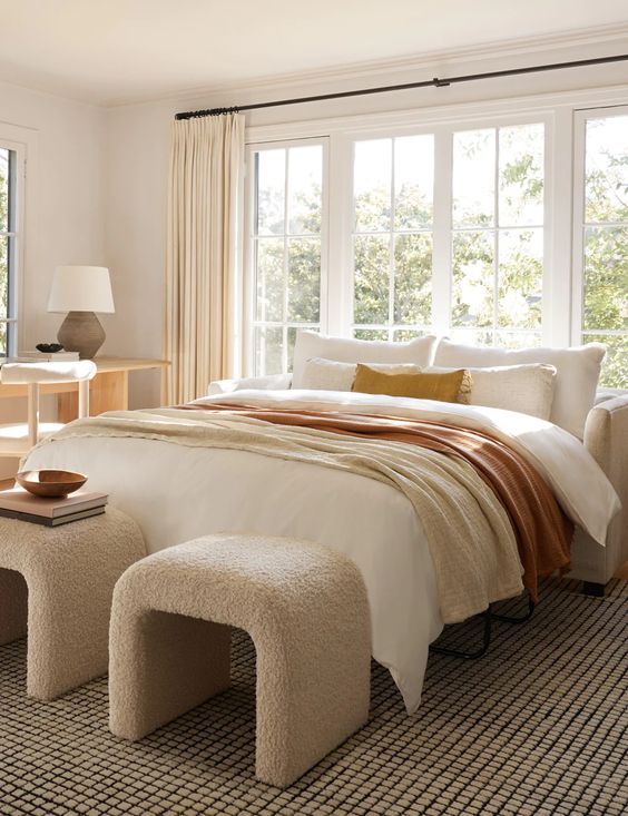 a light-filled bedroom with a white bed and lovely bedding, white boucle stools, a stained vanity and a white chair