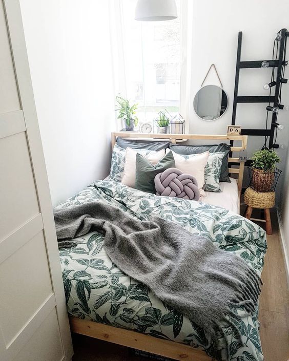 a little narrow bedroom with a stained bed and printed bedding, a nightstand with a plant, a ladder with lights and a mirror