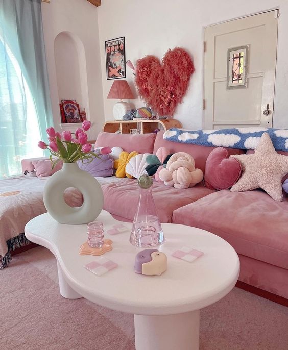 a living room with a pink sectional, colorful pillows, aqua curtains, a heart and some art and a table with pretty decor