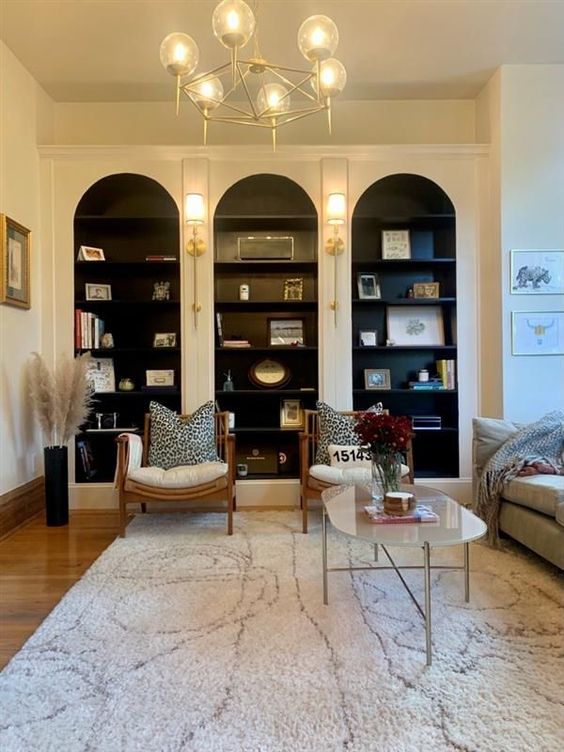 a living room with white arched bookcases with black shelves and backing, white chairs, a neutral sofa, a coffee table and a white rug