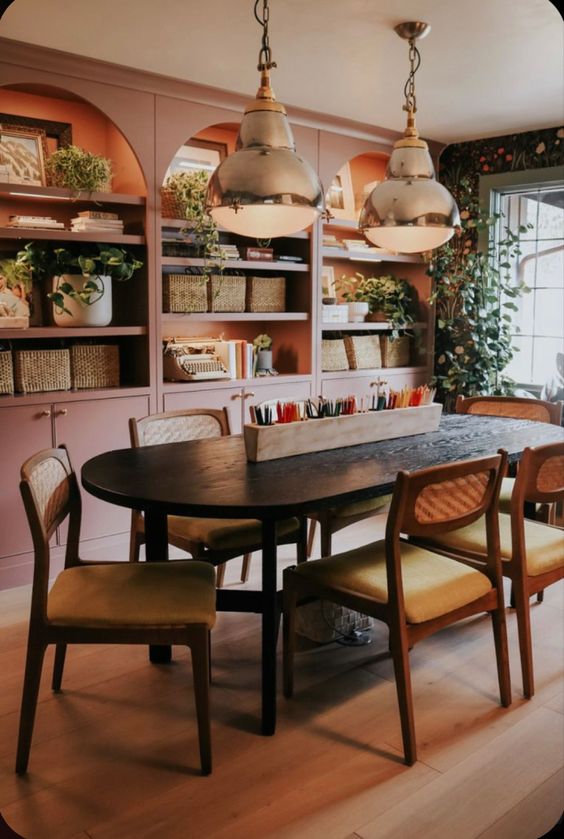 A lovely dining space and home office with a row of built in pink arched bookcases with built in lights, greenery and baskets, a black table and chairs