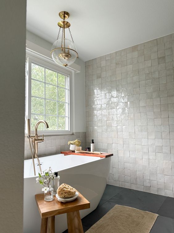 a lovely modern bathroom with a neutral Zellige tile floor, a grey floor, a tub, a wooden stool and a pendant lamp