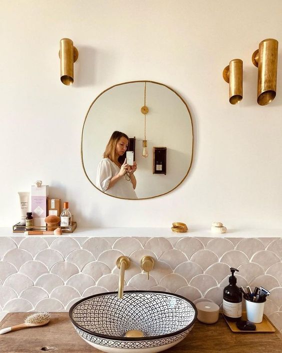 a lovely neutral bathroom clad with grey fish scale tiles, a printed sink, brass tube lamps and fixtures