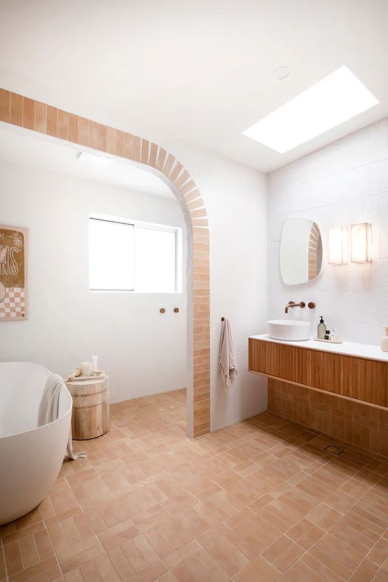 a modern Mediterranean bathroom with a tub, a window, a fluted vanity, a skylight and terracotta tile on the floor