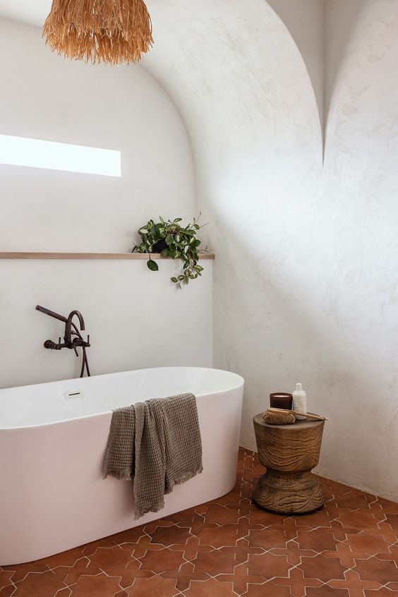 a modern Mediterranean bathroom with an oval tub, a terracotta tile floor, a ledge with greenery and a pendant lamp