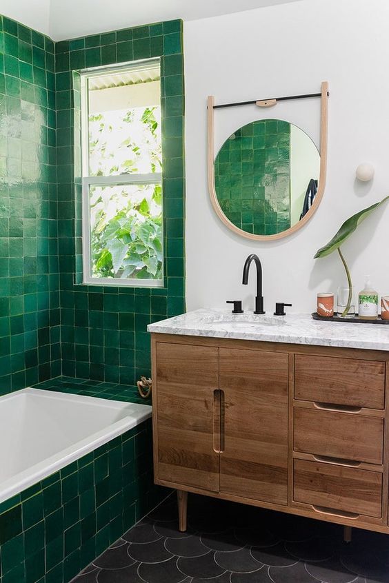a modern bathroom clad with green Zellige tiles, grey fish scale tiles, a stained vanity, a round mirror and some greenery