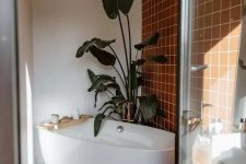 a modern bathroom with an accent wall clad with terracotta tiles, an oval tub, a shower space, a statement plant and a wooden stool