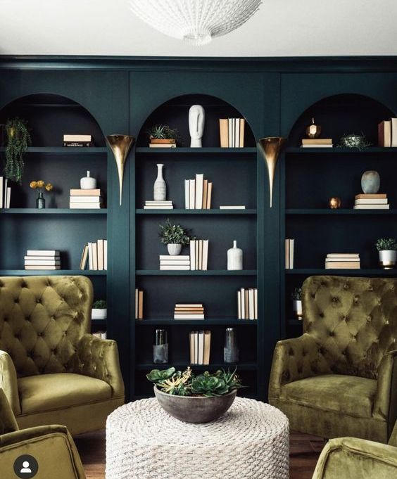 a moody space with dark green arched bookcases, light green tufted chairs and a woven table is very sophisticated