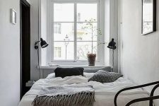 a narrow Scandinavian bedroom with a bed, black sconces and a black chair, some greenery and an artwork