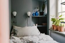 a narrow bedroom in greys, built-in shelves, potted plants and blooms, a bed with neutral bedding and sconces